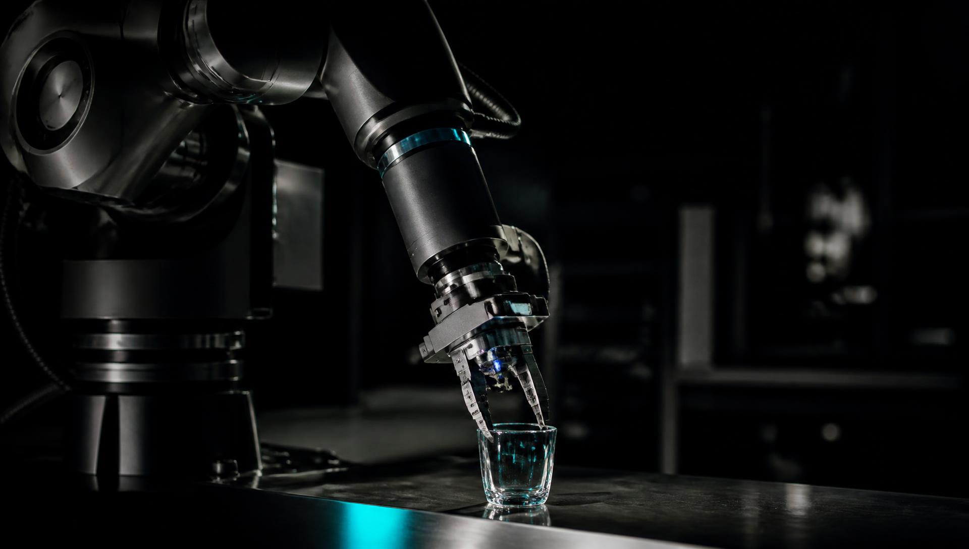 Industrial robotic arm with exposed joints placing a glass object onto a steel surface in a dark robotics laboratory